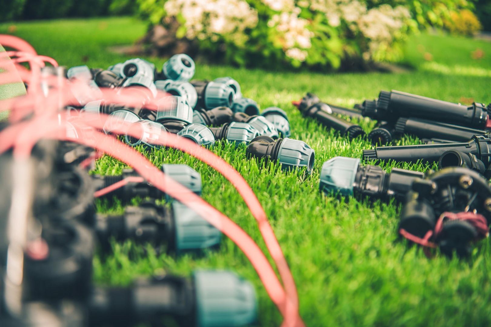 Various irrigation components are arranged on a lush green lawn, ready for installation. Bright sunlight enhances the vibrant colors of the garden surroundings.